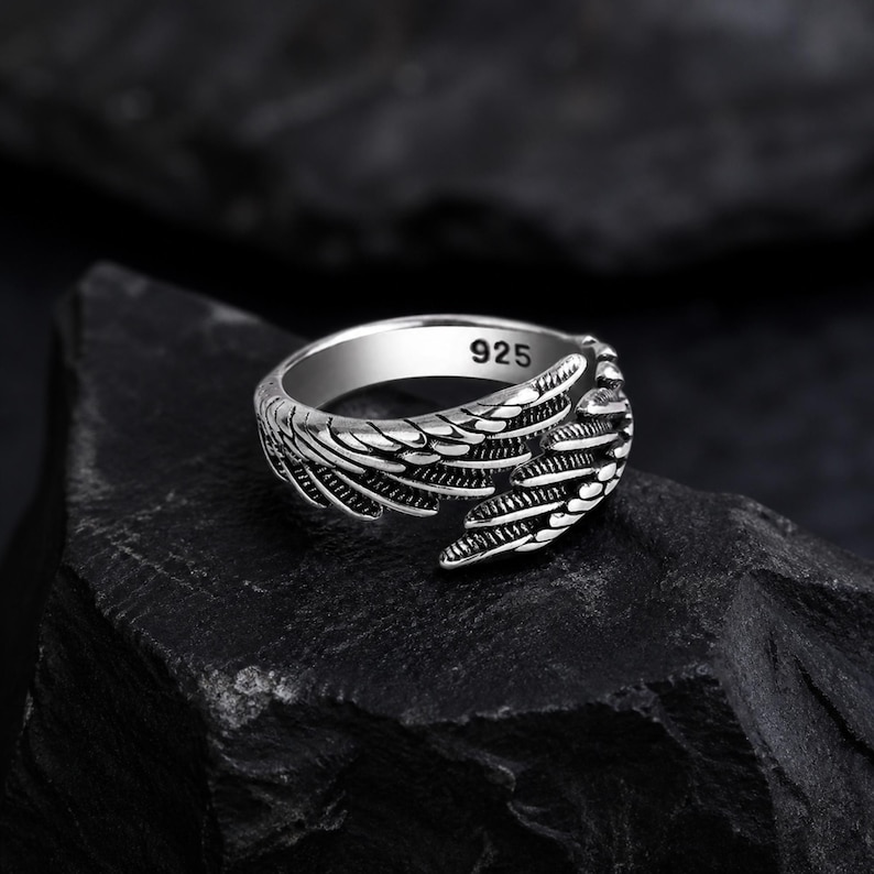 May include: A silver ring with a wing design. The ring is made of polished metal and features intricate feather detailing. The ring is marked with "925" on the inside. The ring is displayed on a dark, textured surface.