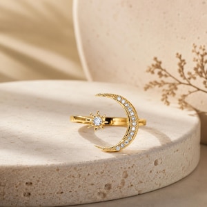 May include: A gold-toned open ring featuring a crescent moon motif on one end and a star motif on the other. The crescent moon is embellished with a row of small clear stones, while the star features a single central clear stone.