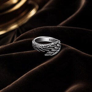 May include: A silver ring with a wing design. The ring is made of a shiny metal and features intricate feather detailing. The number "925" is visible on the ring. The ring is displayed on a dark brown, textured surface.