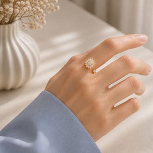 May include: A gold ring with a sunflower design, set with small, clear stones. The ring is on a hand, with a blue sleeve visible. The sunflower has a central cluster of stones and a small dangling charm. The ring is a piece of jewellery.