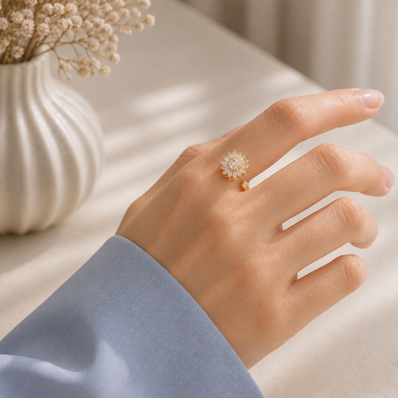 May include: A gold ring with a sunflower design, set with small, clear stones. The ring is on a hand, with a blue sleeve visible. The sunflower has a central cluster of stones and a small dangling charm. The ring is a piece of jewellery.