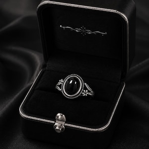 May include: A silver ring with an oval black stone, set in a decorative floral design. The ring is displayed in an open black jewelry box with a silver clasp, against a dark background.