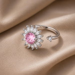 May include: A silver ring with a central pink gemstone encircled by white, petal-shaped stones. The open-design ring has a small, clear stone on the opposite end of the band. The ring is displayed against a neutral, tan-coloured background.