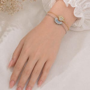 May include: Two delicate bracelets, one gold and one silver, adorn a wrist. The gold bracelet features a sun charm, while the silver one showcases a crescent moon. Both charms are embellished with sparkling crystals, creating a celestial theme.