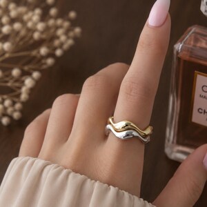 May include: A two-tone ring featuring a stacked design with one smooth gold-coloured band and one smooth silver-coloured band. Both bands have a distinct, undulating wave-like shape that creates a layered appearance when worn together on a finger.