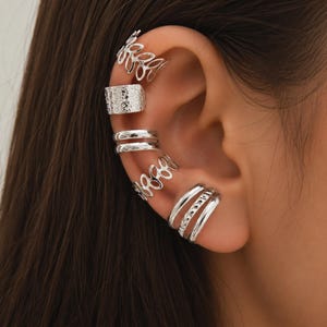Op de afbeelding: Een set van vijf zilverkleurige oorklemmen gedragen langs het kraakbeen. De collectie bevat een klem met bladpatroon, een getextureerde band, een dubbele ringklem, nog een bladpatroonontwerp en een drievoudige bandklem met textuur.