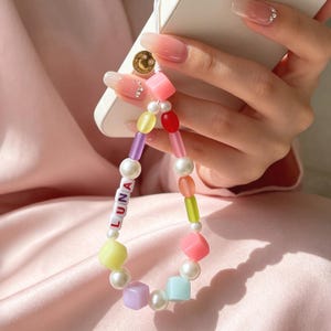 Puede incluir: Un colorido dije para teléfono con el nombre "LUNA" escrito en letras blancas. El dije presenta una variedad de cuentas, incluyendo perlas, cubos y cuentas cilíndricas en colores pastel. El dije está adjunto a un teléfono inteligente blanco.