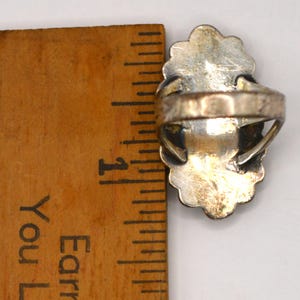 Puede incluir: Un anillo de plata con un engaste de borde festoneado colocado junto a una regla de madera, mostrando que la cara del anillo mide aproximadamente 2,5 cm de largo. La banda de metal est&aacute; unida a la parte posterior del engaste de plata ovalado.