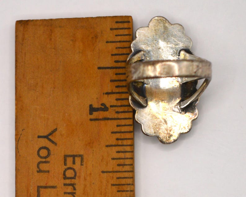 Puede incluir: Un anillo de plata con un engaste de borde festoneado colocado junto a una regla de madera, mostrando que la cara del anillo mide aproximadamente 2,5 cm de largo. La banda de metal est&aacute; unida a la parte posterior del engaste de plata ovalado.
