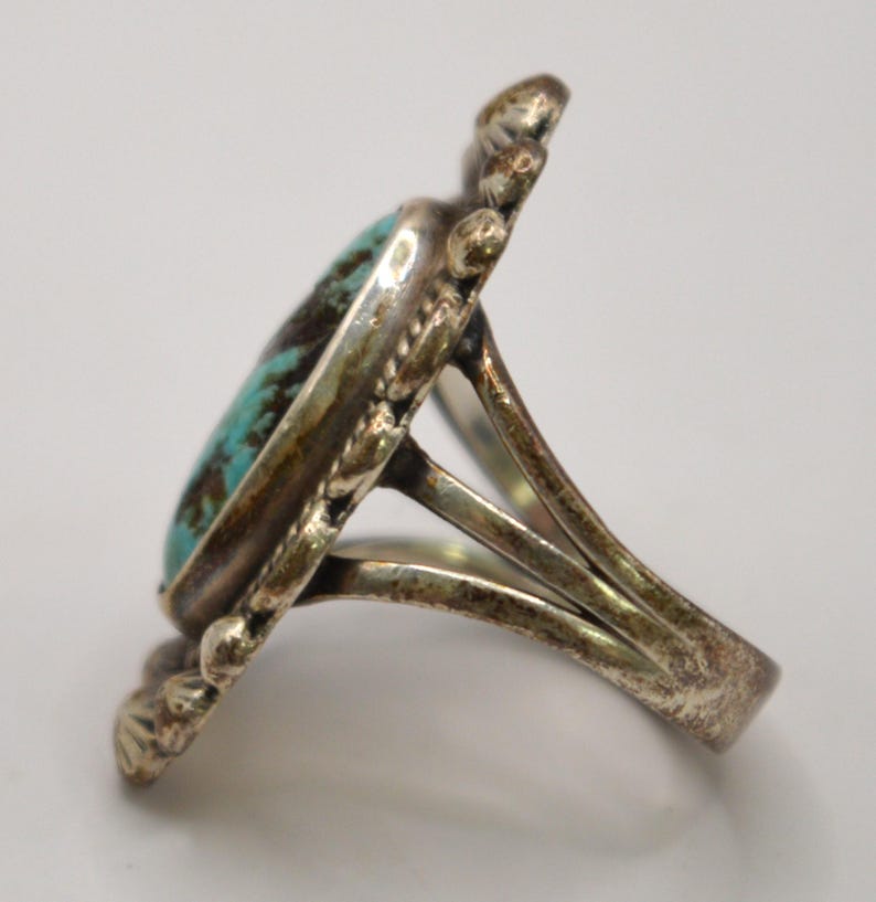 Puede incluir: Un anillo de plata con una piedra turquesa ovalada con patrones de matriz de color marr&oacute;n oscuro. El engaste de metal tiene un borde festoneado decorativo con peque&ntilde;os detalles de cuentas. La banda se divide en tres secciones distintas.