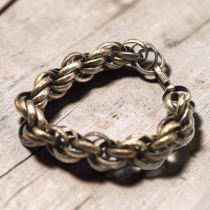 May include: A silver-toned bracelet with a braided chain design. The bracelet is made of interlocking oval links, creating a textured appearance. The clasp is a simple hook and eye closure. The jewelry is displayed on a weathered wooden surface.