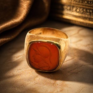 May include: A gold ring with a rectangular, orange-red stone. The stone features a carved profile of a person. The ring has a thick band and a polished finish, showcasing a classic design.