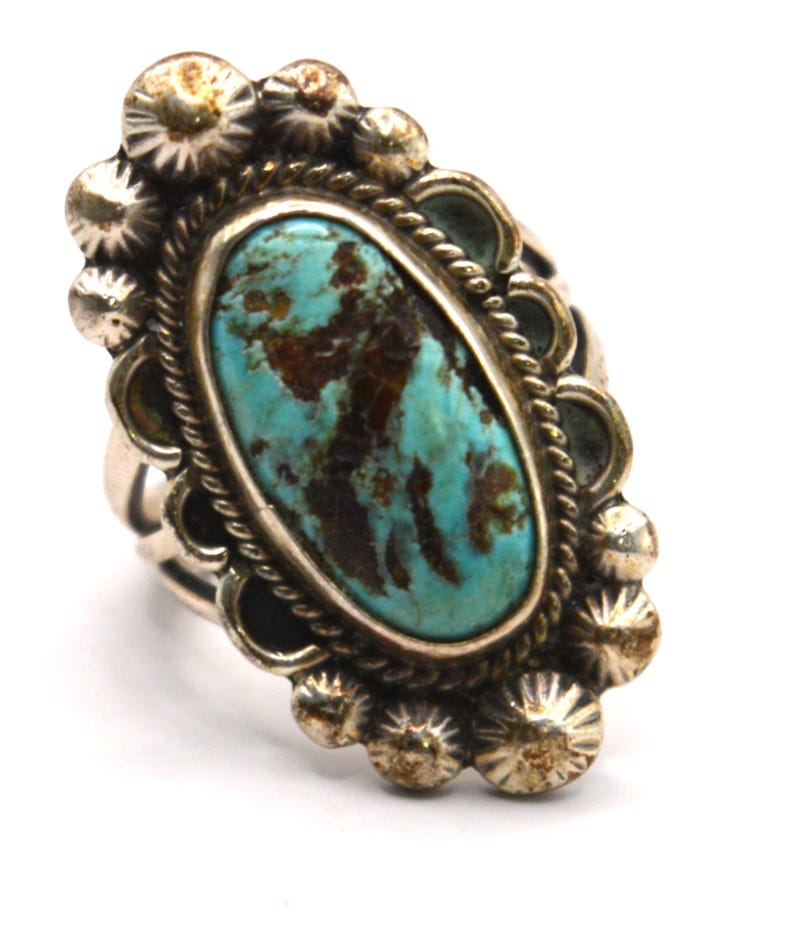 Puede incluir: Un anillo de plata con una gran piedra turquesa de forma ovalada con patrones de matriz de color marr&oacute;n oscuro. La piedra est&aacute; rodeada por un borde de cuerda trenzada y un marco de plata festoneado con peque&ntilde;as cuentas de plata texturizadas.