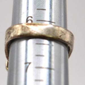 Puede incluir: Un anillo met&aacute;lico con una superficie texturizada e irregular colocado en un mandril de joyer&iacute;a plateado. El anillo descansa entre las marcas de tama&ntilde;o 6 y 7 de la herramienta, indicando un di&aacute;metro de aproximadamente 16,9 mm en la escala.