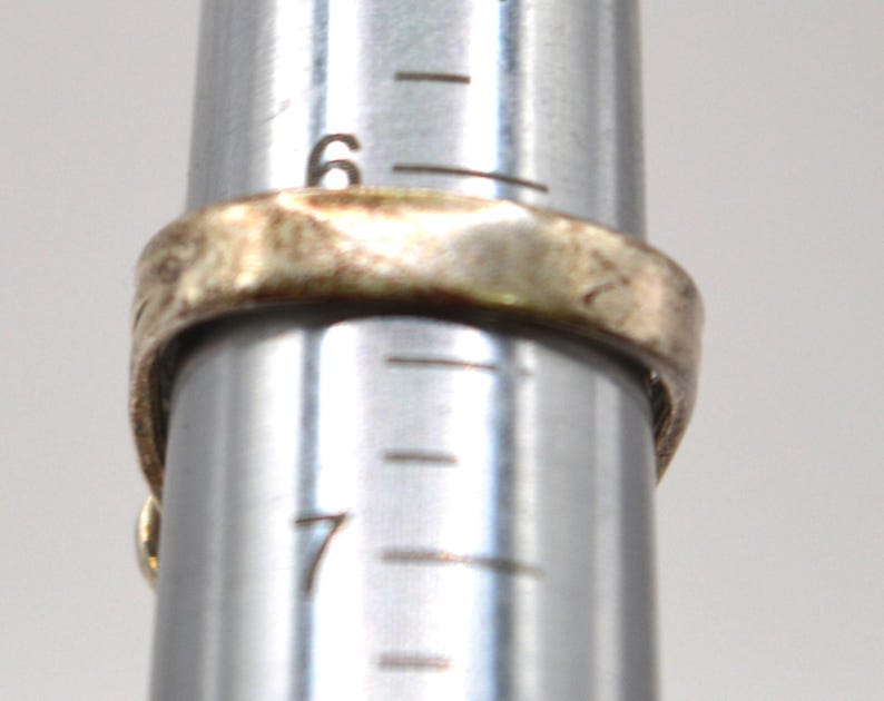 Puede incluir: Un anillo met&aacute;lico con una superficie texturizada e irregular colocado en un mandril de joyer&iacute;a plateado. El anillo descansa entre las marcas de tama&ntilde;o 6 y 7 de la herramienta, indicando un di&aacute;metro de aproximadamente 16,9 mm en la escala.