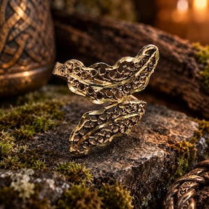 May include: A gold-toned metal ring featuring two overlapping leaf-shaped elements with an intricate, openwork filigree pattern. The surface of the leaves has a textured, porous appearance, and the band is open, allowing for adjustment.