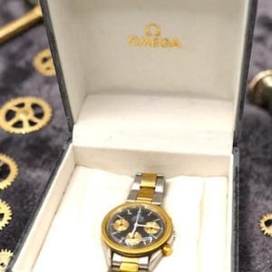 May include: A vintage Omega watch with a black dial and gold accents, presented in its original gray box. The watch features a gold bezel and a two-tone gold and silver band. The box is lined with white fabric, and the Omega logo is visible.