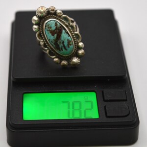 Puede incluir: Un anillo con piedra turquesa ovalada engastada en un marco de plata con detalles decorativos de cuentas y alambre trenzado. El anillo est&aacute; sobre una b&aacute;scula digital negra que muestra un peso de 7,82 gramos en la pantalla verde retroiluminada.