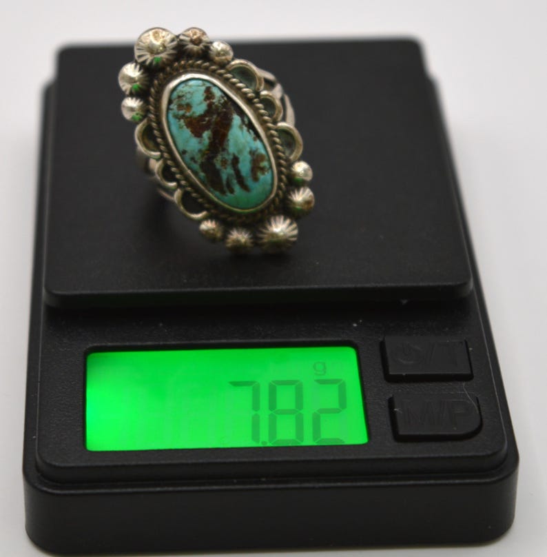 Puede incluir: Un anillo con piedra turquesa ovalada engastada en un marco de plata con detalles decorativos de cuentas y alambre trenzado. El anillo est&aacute; sobre una b&aacute;scula digital negra que muestra un peso de 7,82 gramos en la pantalla verde retroiluminada.
