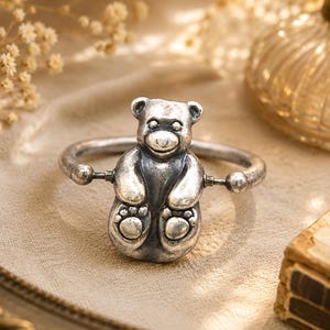 May include: A silver-toned metal bangle bracelet featuring a central three-dimensional teddy bear charm. The bear has a smooth, polished finish with detailed paws and facial features. The circular band connects to the sides of the bear figure.