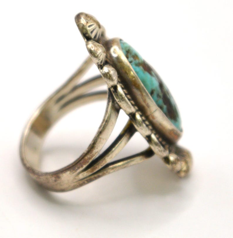 Puede incluir: Un anillo de plata con una piedra turquesa ovalada con patrones de matriz de color marr&oacute;n oscuro. La banda de metal est&aacute; dividida en tres secciones y la piedra est&aacute; rodeada por un borde de plata decorativo con peque&ntilde;os acentos met&aacute;licos redondeados.