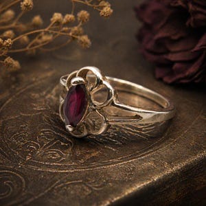 May include: A silver ring with an ornate, openwork design. The ring features a marquise-cut, deep red gemstone at its center. The ring is set against a dark, textured background, highlighting the intricate details of the jewelry.