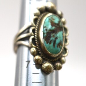 Puede incluir: Un anillo de plata con una piedra turquesa ovalada con matriz marr&oacute;n, rodeada por un borde decorativo de cuentas. El anillo est&aacute; colocado en un mandril de metal, con la banda descansando cerca de la marca 7 en la herramienta de medici&oacute;n.