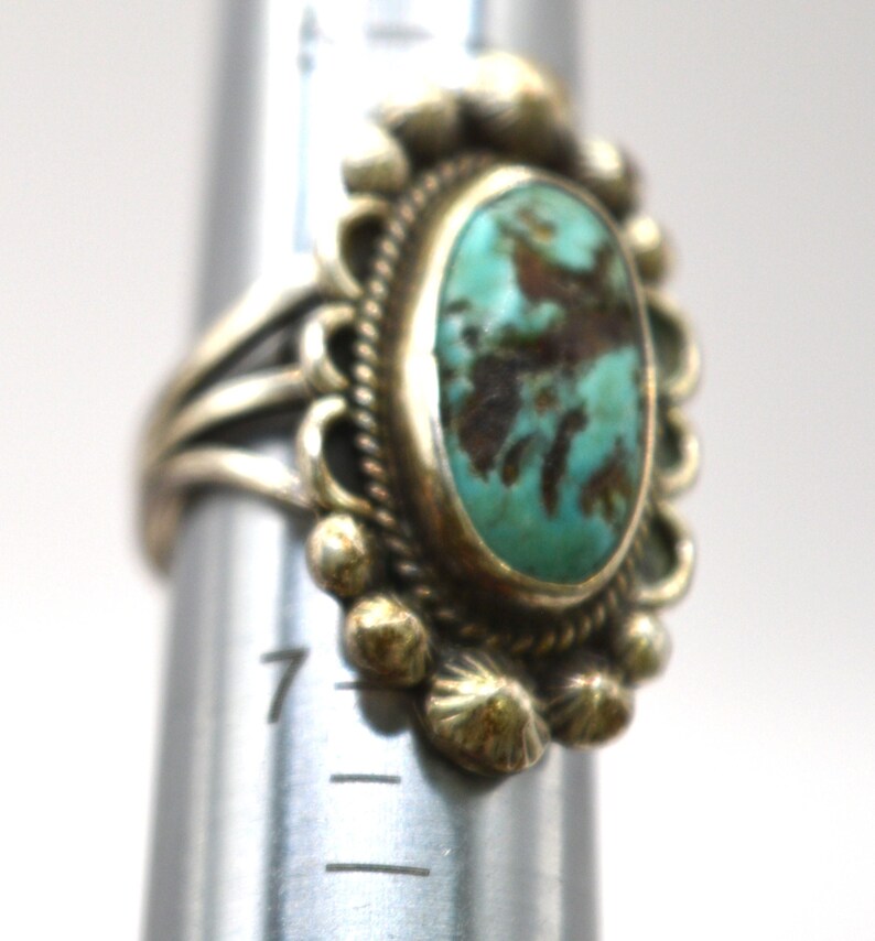 Puede incluir: Un anillo de plata con una piedra turquesa ovalada con matriz marr&oacute;n, rodeada por un borde decorativo de cuentas. El anillo est&aacute; colocado en un mandril de metal, con la banda descansando cerca de la marca 7 en la herramienta de medici&oacute;n.