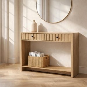 May include: A light wood console table with two drawers and a lower shelf. The drawers have vertical grooved detailing and black knobs. A woven basket sits on the lower shelf, and a beige vase is on top. A round gold mirror hangs above.