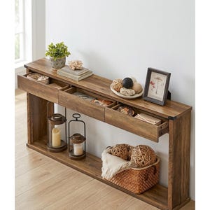 May include: A wooden console table with three drawers, displaying decorative items. The table features a potted plant, books, a framed picture, and a woven basket with decorative balls. Two candle lanterns are placed below the drawers.