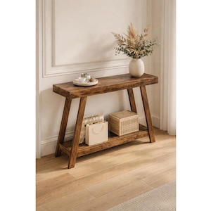 May include: A rustic, wooden console table with a lower shelf. The table is a warm brown color and has a natural wood grain. A white vase with dried flowers sits on top, along with a tray of small items. A woven basket and shopping bags are on the lower shelf.
