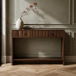 May include: A rectangular wooden console table featuring a single drawer with vertical slat detailing and two small black knobs. The table includes a lower open shelf and is topped with a light-colored ceramic vase containing a dried plant stem.