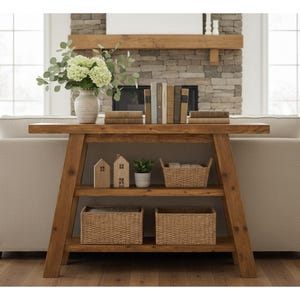 May include: A wooden console table with three shelves, holding decorative items. The top shelf displays books and a vase with flowers. The lower shelves feature woven baskets, small wooden houses, and a potted plant. The table is set against a neutral-toned sofa.