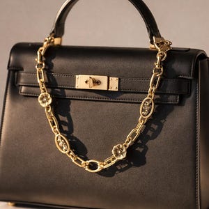 May include: Black leather handbag with a top handle and gold-tone hardware. A decorative gold chain with floral and oval embellishments hangs from the bag. The bag has a structured shape and a clasp closure.