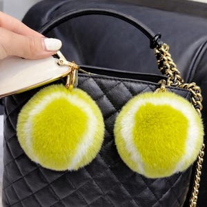 May include: Black quilted handbag with a gold chain strap and a black handle. Two fluffy yellow and white tennis ball-shaped keyrings hang from the bag. The bag is held by a person.