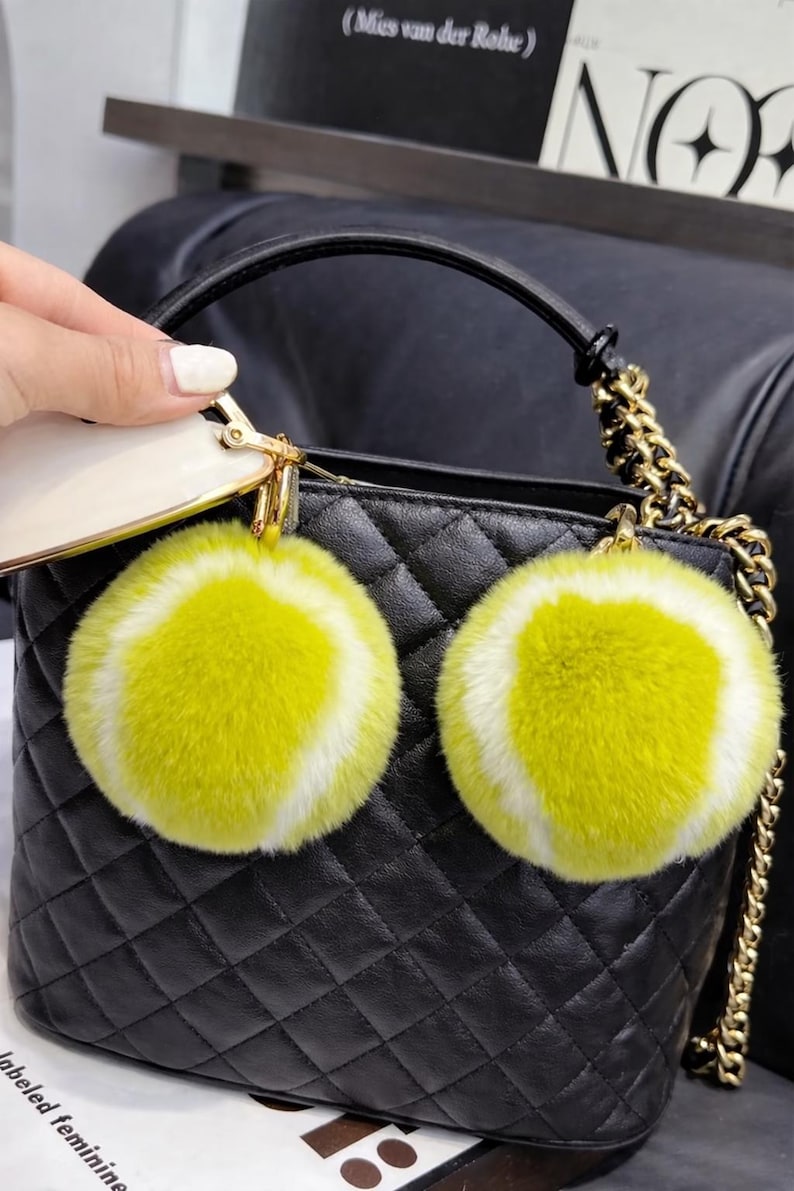 May include: Black quilted handbag with a gold chain strap and a black handle. Two fluffy yellow and white tennis ball-shaped keyrings hang from the bag. The bag is held by a person.