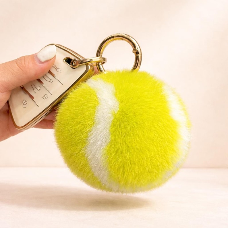 May include: A yellow and white tennis ball-shaped keyring with a gold-coloured clasp and ring. The keyring is attached to a car key with a gold-coloured border.