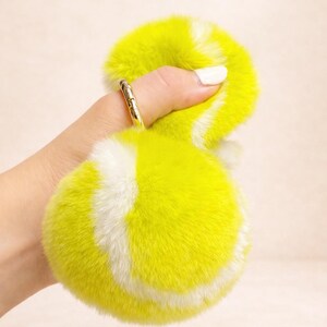 May include: A fluffy, yellow and white tennis ball-shaped keyring. The keyring is made of soft, faux fur material. The ring is gold-coloured.