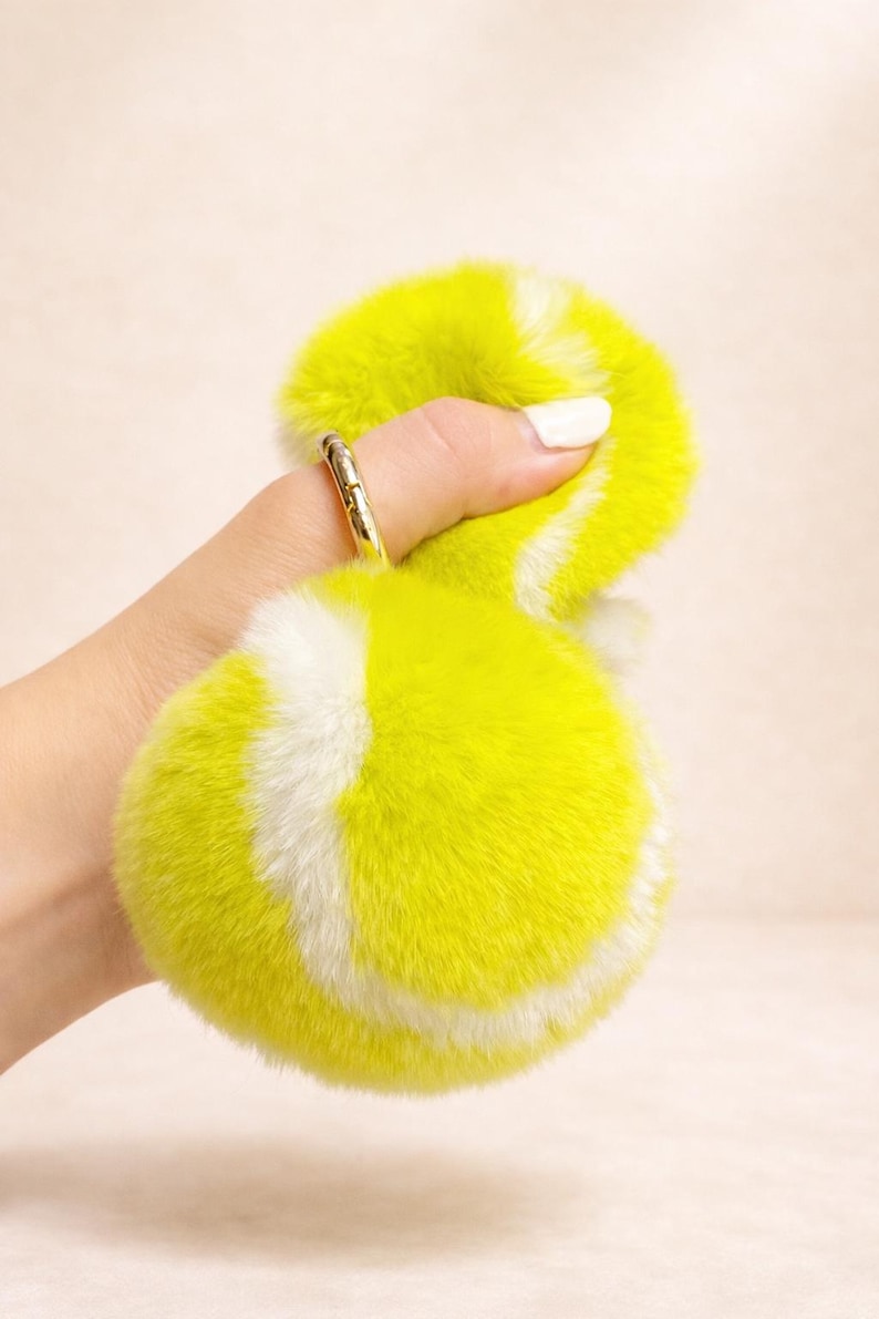 May include: A fluffy, yellow and white tennis ball-shaped keyring. The keyring is made of soft, faux fur material. The ring is gold-coloured.