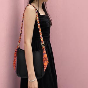 Puede incluir: Bolso de hombro negro con una correa decorativa con detalles en naranja y morado. El bolso tiene un diseño perforado y se combina con una bufanda estampada. La correa es una combinación de cadena dorada y tela.