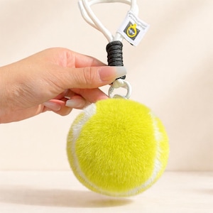 May include: A yellow tennis ball-shaped pet toy with white stripes and a white rope handle. The toy has a black and white accent near the top and a small tag attached to the handle.
