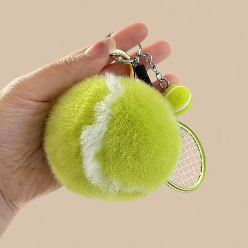 May include: A tennis-themed keyring featuring a large, fluffy green tennis ball with a white stripe, a small tennis ball, and a miniature tennis racket. The keyring is attached to a silver metal ring and chain.