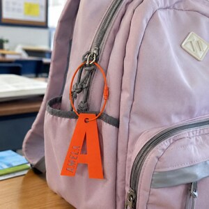 Könnte beinhalten: Ein leuchtend orangefarbener Kofferanhänger in Form des Großbuchstabens A, auf dessen linker Seite der Name AMELA vertikal eingeprägt ist. Der Anhänger ist mit einer dünnen, kreisförmigen orangefarbenen Drahtschlaufe am Reißverschluss eines helllila Rucksacks befestigt.