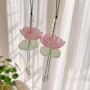 Könnte beinhalten: Zwei dekorative Hängeornamente mit rosa Lotusblüten und grünen Seerosenblättern. Jedes Ornament hat eine Kette aus klaren Perlen und eine dunkelgrüne Kordel. Der Hintergrund ist ein weißer Vorhang.