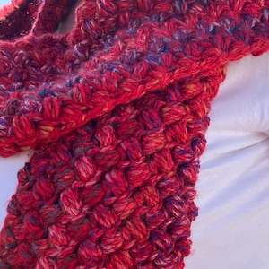 May include: A hand-knitted red scarf with a textured, bubble-like pattern. The scarf is made of thick yarn with flecks of blue, yellow, and purple. The scarf is wrapped around the neck.