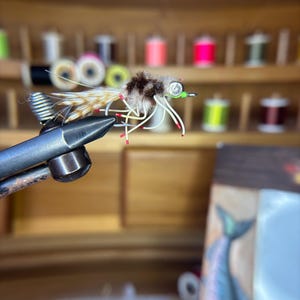 May include: A close-up of a handmade fishing fly. The fly features a brown and tan body with white and red accents. It is held in a metal vise, with a wooden storage box and spools of thread in the background.