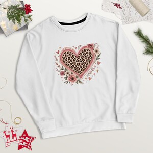 Peut inclure: Un sweat-shirt blanc à manches longues arborant un motif central en forme de cœur avec un imprimé léopard, entouré de roses roses, de feuilles vertes et de petits cœurs et étoiles dispersés sur le devant du vêtement.