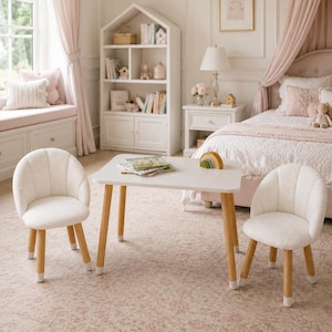 Op de afbeelding: Een witte rechthoekige tafel met vier licht houten poten en twee bijpassende witte stoelen met afgeronde, schelpvormige rugleuningen en licht houten poten. Op de tafel liggen een klein houten regenboogspeeltje en een stapel boeken.