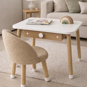 May include: A white children's table with a tan drawer and wooden legs, paired with a matching chair. A wooden rainbow and a book rest on the table. The chair and table are on a light-coloured rug.