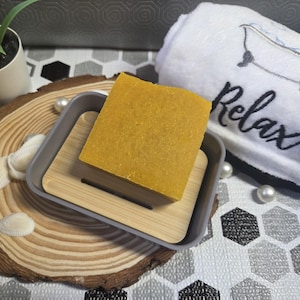 May include: A yellow soap bar sits on a grey and bamboo soap dish, on a wooden slice with seashells. A white towel with the word "Relax" embroidered is in the background, along with a small potted plant.
